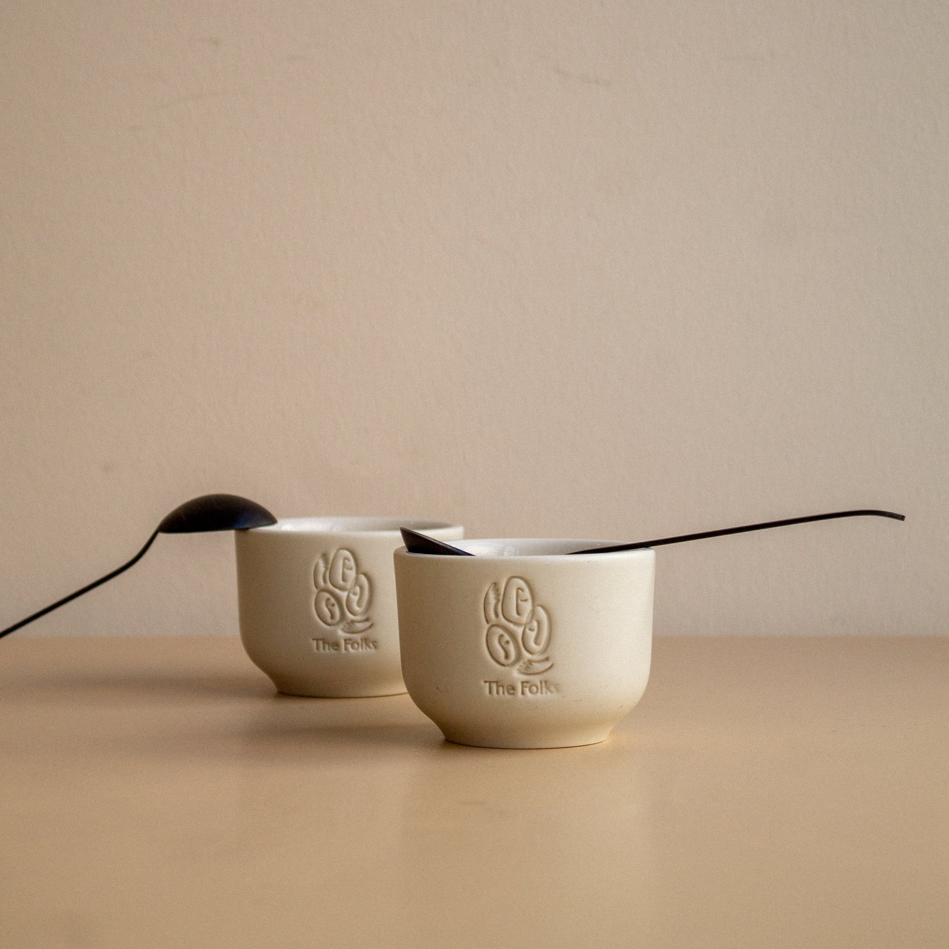 Ceramic mug featuring The Folks logo next to a Comandante coffee grinder on a wooden surface.